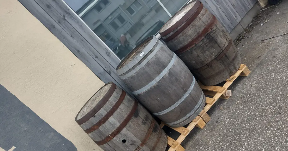 Drei Holzfässer mit Metallbändern stehen auf einer Holzpalette vor einem Gebäude mit Holzverkleidung im rheinischen Stil und einem Fenster. Bierkultur im Rheinland.