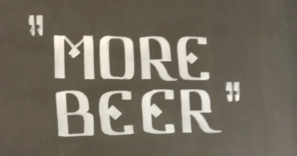 Der Text an der Wand lautet: "MORE BEER" (Mehr Bier) in großen, stilisierten weißen Buchstaben mit Anführungszeichen am Anfang und am Ende - eine Anspielung auf alle Craftbier-Fans.