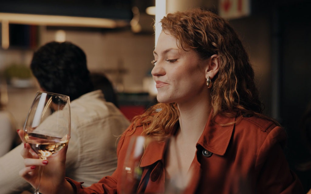 Eine Frau mit lockigem Haar in einem roten Mantel sitzt mit einem Glas Weißwein in der Hand an einem Tisch in einem schwach beleuchteten Restaurant.
