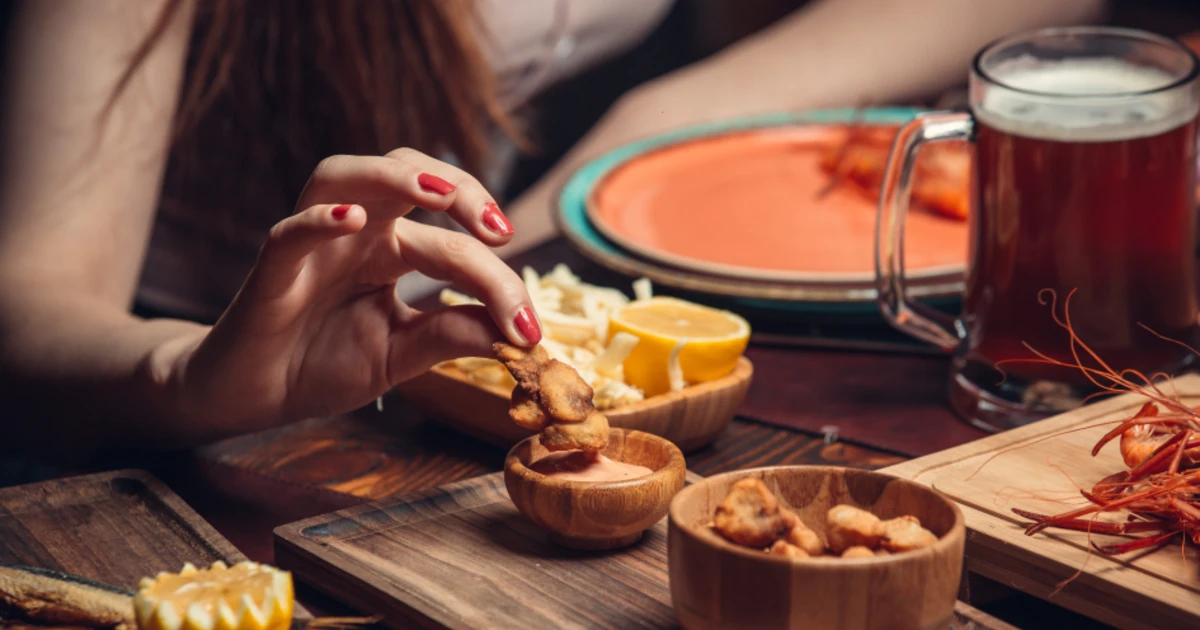 Eine Person mit rotem Nagellack nimmt einen Snack aus einer Holzschale, umgeben von Bier, Pairing mit Zitronenspalten, Käse und Garnelen - eine einladende Geschmackspaarung auf dem Tisch.