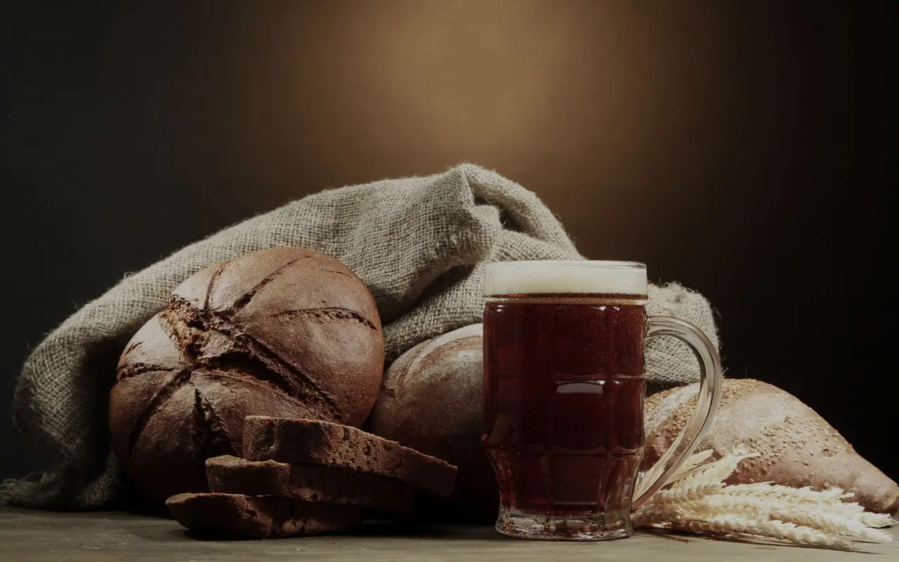 Ein Glaskrug mit dunklem Bier steht neben runden, teilweise aufgeschnittenen Schwarzbrotlaiben, im Hintergrund ein Leinensack und Weizenhalme.